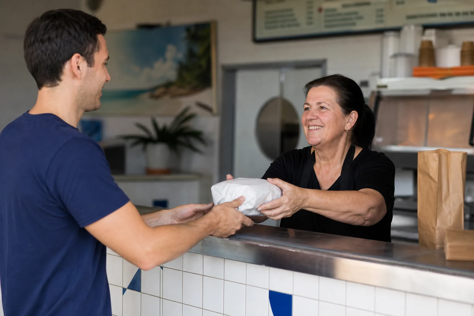 A takeaway shop worker handing a wrapped food order to a customer at the counter
